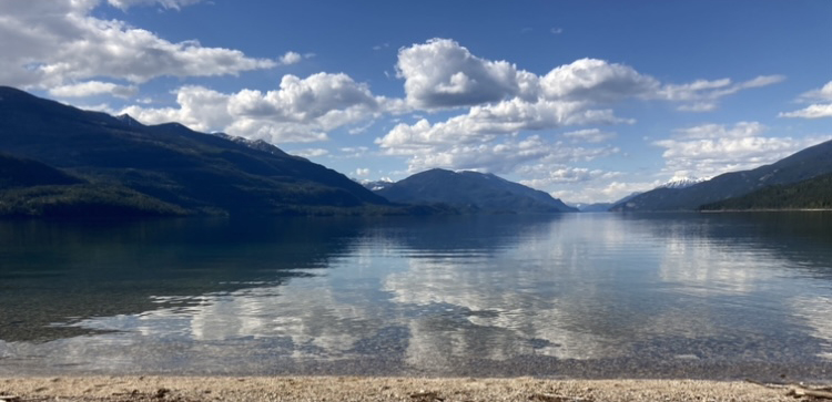 Photo of Kootenay Lake under a blue sky with fluffy white clouds. The mountains depend to the edges of the river, and the water is very still, reflecting the mountains and sky. A narrow strip of beach is showing in the foreground.